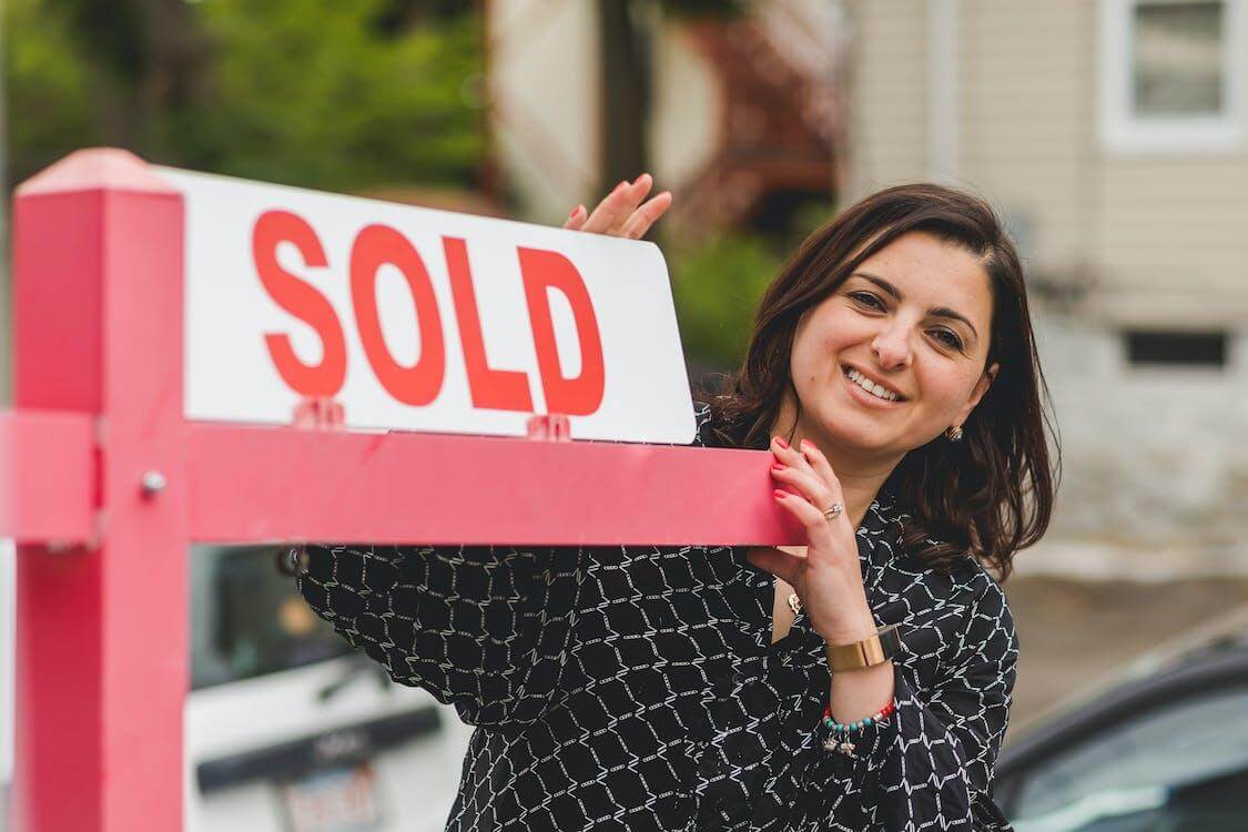 Free Real Estate Agent Smiling in Black Printed Blouse Holding a Sold Signage Metal Post Outside a House Stock Photo
